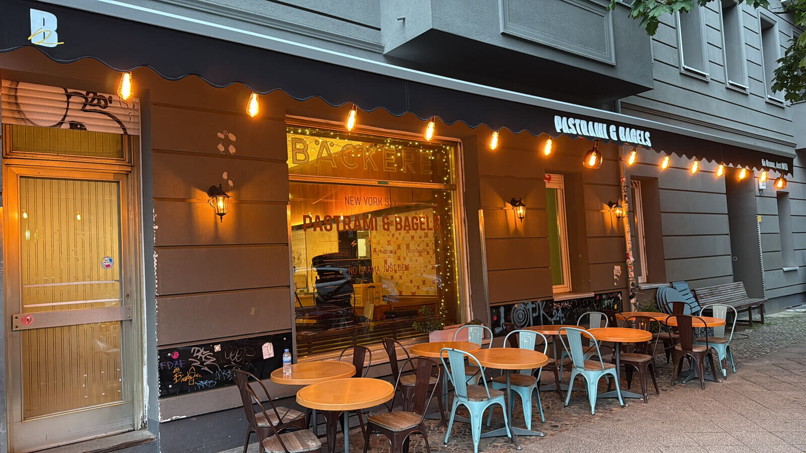 Börk's Deli in Berlin am Abend — Außenbestuhlung, Lichterkette und Pastrami & Bagels Markise